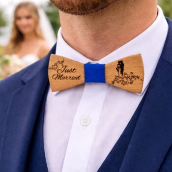 Κοντινή λήψη γαμπρού με μπλε κοστούμι και λευκό πουκάμισο, που φοράει ξύλινο παπιγιόν με χάραξη “Just Married” και μπλε λεπτομέρεια στο κέντρο, σε περιβάλλον γάμου, με τη νύφη θολή στο φόντο.