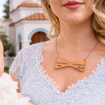 Κοντινή λήψη γυναίκας που κρατά μωρό ντυμένο με λευκό βαπτιστικό φόρεμα σε τελετή βάπτισης. Η γυναίκα φορά ανοιχτό γαλάζιο δαντελένιο φόρεμα και ξύλινο κολιέ σε σχήμα παπιγιόν, χαραγμένο με ημερομηνία και όνομα. Το φόντο είναι απαλά θολό και παραπέμπει σε εκκλησία, με φυσικό φως και ήρεμη, τελετουργική ατμόσφαιρα.