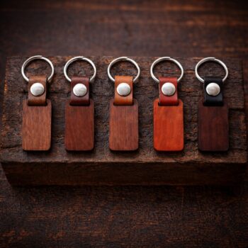 Πέντε ξύλινα μπρελόκ τοποθετημένα δίπλα-δίπλα πάνω σε σκούρο, rustic ξύλινο μπλοκ, με μεταλλικό κρίκο και δερμάτινο λουράκι σε πέντε χρώματα: καρυδί, μαόνι, αγγλικό φυσικό, κόκκινο και μαύρο, σε ατμοσφαιρικό φωτισμό χωρίς χάραξη στο ξύλο.