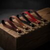 : Five leather keychains in mahogany, walnut, black, red, and dark English bridle brown are lined up on a rustic wood block.