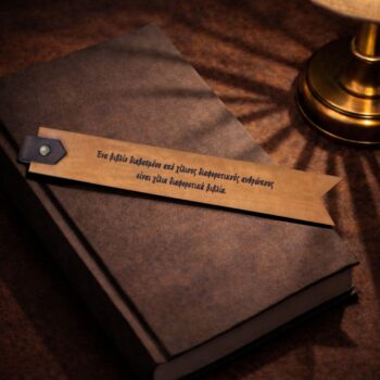 Κοντός ταμπά δερμάτινος σελιδοδείκτης με χαραγμένο ελληνικό κείμενο, τοποθετημένος πάνω σε κλειστό βιβλίο με γκρι εξώφυλλο. Στο επάνω μέρος υπάρχει μικρή δερμάτινη λεπτομέρεια με μεταλλικό κουμπί. Το φως από επιτραπέζιο φωτιστικό δημιουργεί απαλές σκιές, δίνοντας ατμοσφαιρική, ζεστή αίσθηση.