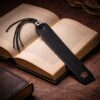 Μαύρος δερμάτινος σελιδοδείκτης με κομμένο αστέρι στο κάτω μέρος, τοποθετημένος πάνω σε ανοιχτό παλιό βιβλίο, με μαύρα δερμάτινα κορδόνια και μεταλλική λεπτομέρεια στο επάνω μέρος, φωτογραφημένος σε ατμοσφαιρικό, ζεστό φωτισμό με απαλές σκιές.