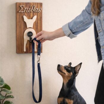 Ξύλινη κρεμάστρα λουριού σκύλου με χαραγμένο όνομα στον τοίχο. Λουρί κρέμεται στο άγκιστρο, ενώ ένας μαύρος και καφέ pinscher κάθεται από κάτω, περιμένοντας να βγει βόλτα, με γυναικείο χέρι να πλησιάζει το λουρί.