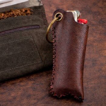 An open dark green suede tobacco pouch rests on a rustic wooden table, showing loose tobacco and a single cigarette filter placed on rolling paper. Beside it stands a handcrafted brown leather lighter cover holding a red Bic lighter, with warm, moody lighting and soft shadows highlighting the textures of the leather and wood.
