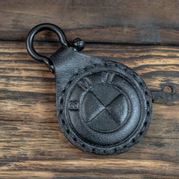 Ένα μικρό, στρογγυλό δερμάτινο μπρελόκ σε μαύρο χρώμα. Στο κέντρο έχει ανάγλυφο ένα κυκλικό σχήμα που θυμίζει το στυλιζαρισμένο λογότυπο της BMW, με τα γράμματα B, M, W χαραγμένα γύρω από τον κύκλο. Η υφή του δέρματος είναι απαλή αλλά εμφανώς ραμμένη περιμετρικά, με μαύρες ραφές. Στο επάνω μέρος υπάρχει ένας μεταλλικός κρίκος σε μαύρο χρώμα για να κρεμιέται σε κλειδιά ή τσάντα. Η συνολική εμφάνιση είναι κομψή και μίνιμαλ, με έμφαση στο ανάγλυφο σχέδιο.