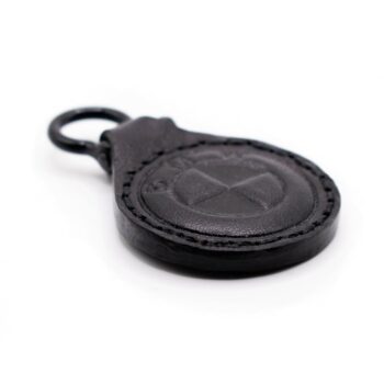 Ένα μικρό, στρογγυλό δερμάτινο μπρελόκ σε μαύρο χρώμα. Στο κέντρο έχει ανάγλυφο ένα κυκλικό σχήμα που θυμίζει το στυλιζαρισμένο λογότυπο της BMW, με τα γράμματα B, M, W χαραγμένα γύρω από τον κύκλο. Η υφή του δέρματος είναι απαλή αλλά εμφανώς ραμμένη περιμετρικά, με μαύρες ραφές. Στο επάνω μέρος υπάρχει ένας μεταλλικός κρίκος σε μαύρο χρώμα για να κρεμιέται σε κλειδιά ή τσάντα. Η συνολική εμφάνιση είναι κομψή και μίνιμαλ, με έμφαση στο ανάγλυφο σχέδιο.