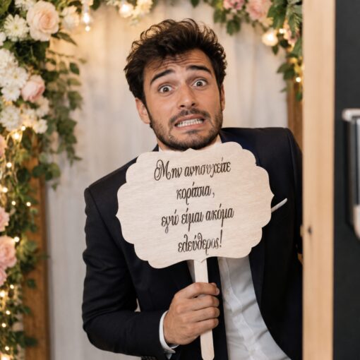 Νεαρός άνδρας κρατά ξύλινο πλακάτ photobooth γάμου με ελληνικό αστείο μήνυμα.