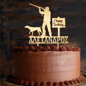 A round chocolate cake covered in smooth chocolate frosting is shown from the side, with decorative piped chocolate swirls along the top and bottom edges. On top of the cake stands a wooden cake topper in a natural light wood color. The topper depicts the silhouette of a hunter aiming a rifle, with a hunting dog standing beside him. Next to the figures, there is a small wooden sign that reads “Happy Birthday” in handwritten-style lettering. Below the figures, the name “ΑΛΕΞΑΝΔΡΟΣ” is cut out in Greek capital letters. The background is dark and softly blurred, making the cake and topper stand out clearly.