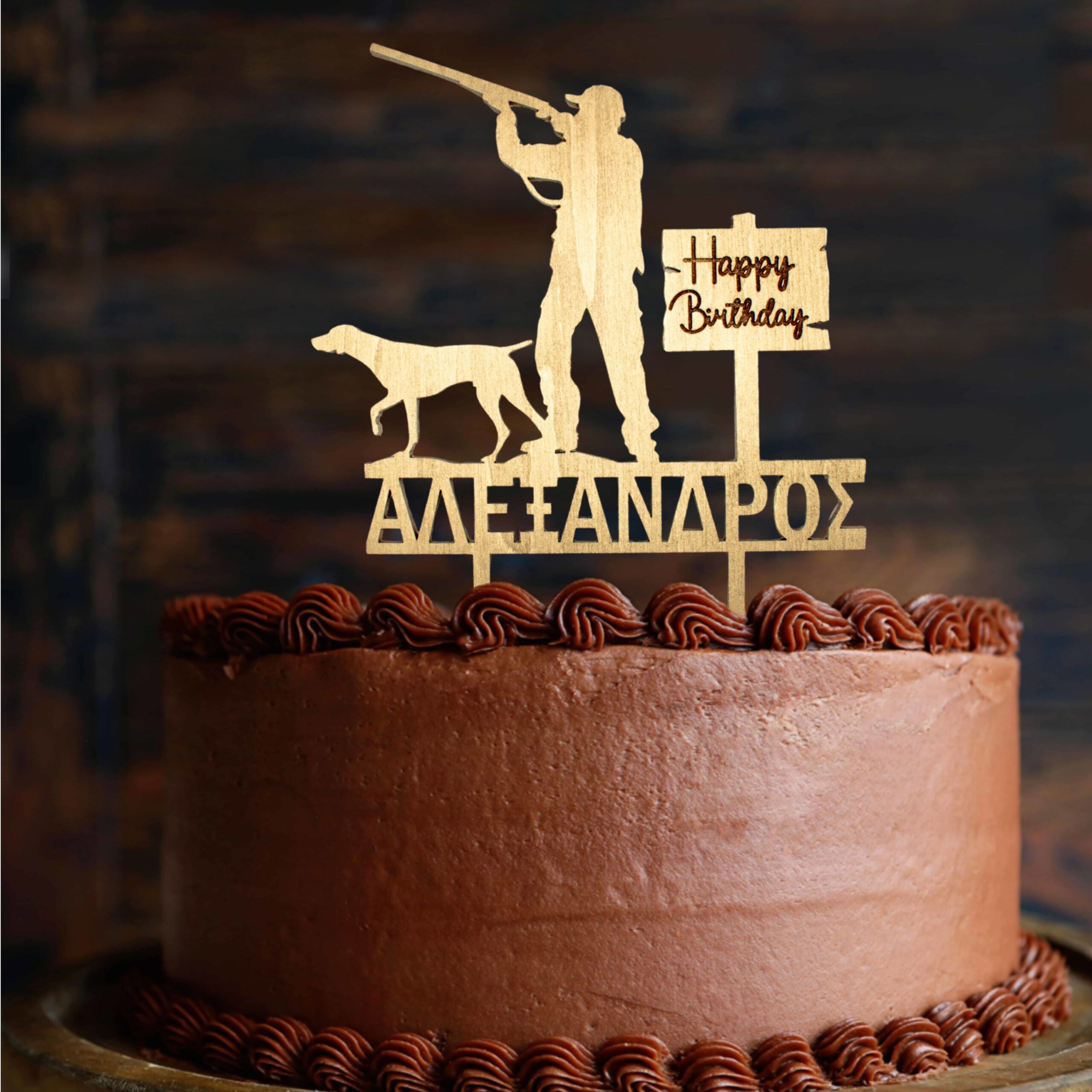 A round chocolate cake covered in smooth chocolate frosting is shown from the side, with decorative piped chocolate swirls along the top and bottom edges. On top of the cake stands a wooden cake topper in a natural light wood color. The topper depicts the silhouette of a hunter aiming a rifle, with a hunting dog standing beside him. Next to the figures, there is a small wooden sign that reads “Happy Birthday” in handwritten-style lettering. Below the figures, the name “ΑΛΕΞΑΝΔΡΟΣ” is cut out in Greek capital letters. The background is dark and softly blurred, making the cake and topper stand out clearly.