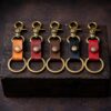 “Five handmade leather keychains in different colors—tan, mahogany, walnut brown, black, and red—lined up side by side on a dark, rustic wooden block. Each keychain features antique brass hardware with a swivel clasp and round key ring, photographed in moody lighting against a dark background.”