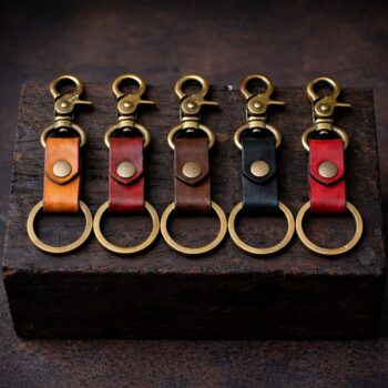 “Five handmade leather keychains in different colors—tan, mahogany, walnut brown, black, and red—lined up side by side on a dark, rustic wooden block. Each keychain features antique brass hardware with a swivel clasp and round key ring, photographed in moody lighting against a dark background.”